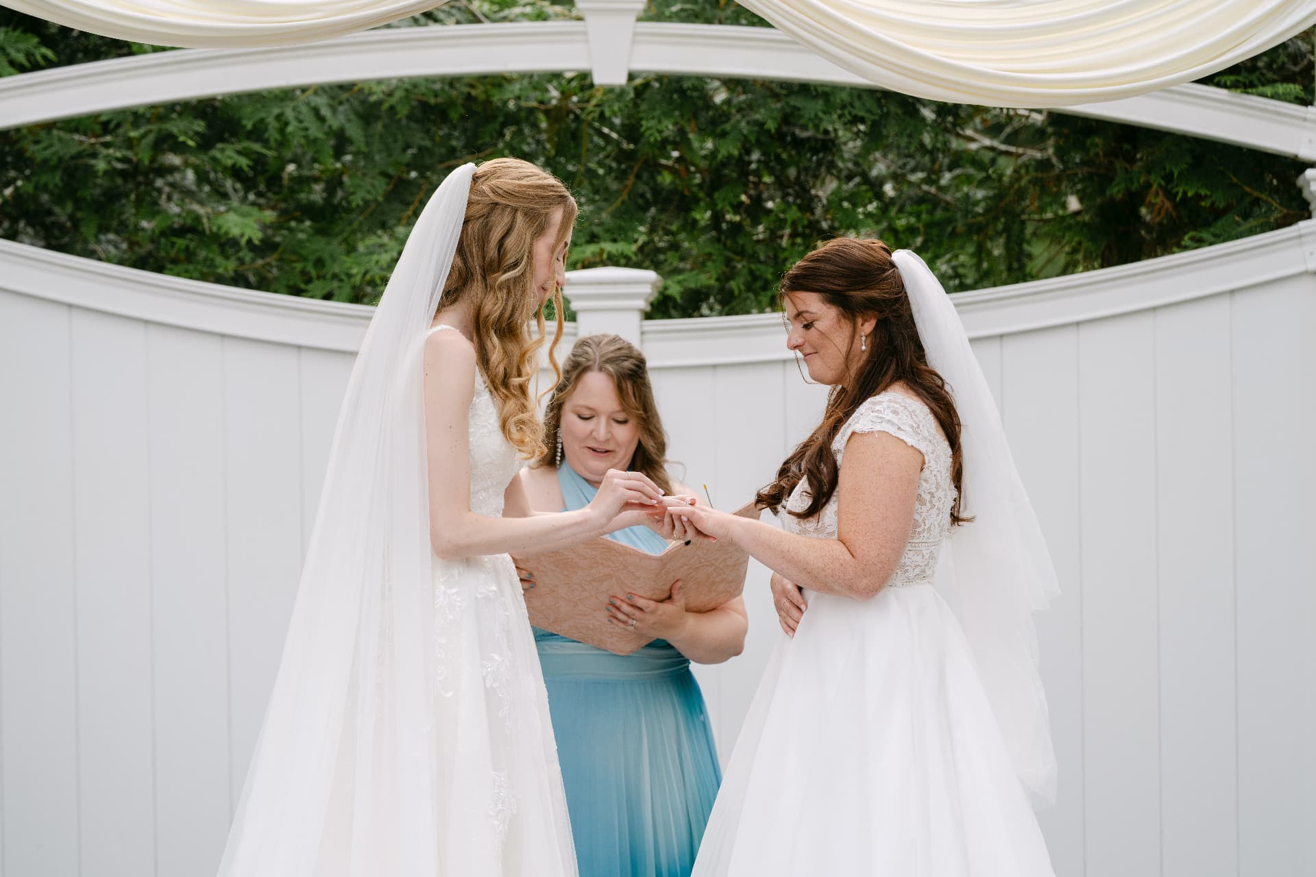Two brides exchanging rings beneath a draped garden gazebo