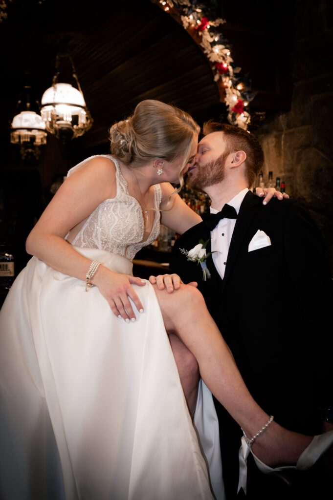 Bride sitting on the groom’s lap while sharing a kiss inside the dimly lit underground lounge at Union Station Banquets. Warm hanging lights and floral accents glow in the background, creating a romantic and intimate wedding portrait with an elegant vintage atmosphere.