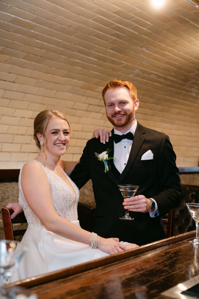 Massachusetts wedding Photographer photographs Bride and groom smiling together inside the historic underground tunnel at Union Station Banquets. The groom holds a martini while the couple leans against polished wood seating beneath arched brick ceilings and warm ambient lighting, creating a relaxed and elegant wedding portrait.