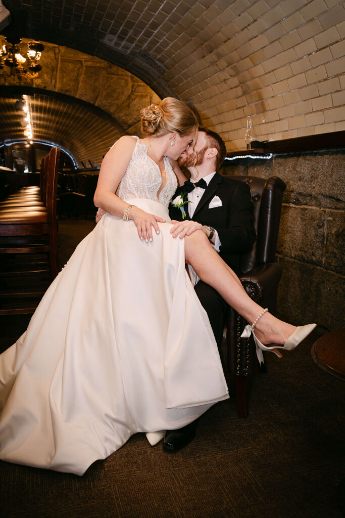 Bride sitting on the groom’s lap while sharing a kiss inside the historic underground tunnel at Union Station Banquets. Warm lighting, arched brick ceilings, and dark leather seating create a romantic and dramatic wedding portrait with an elegant vintage feel.