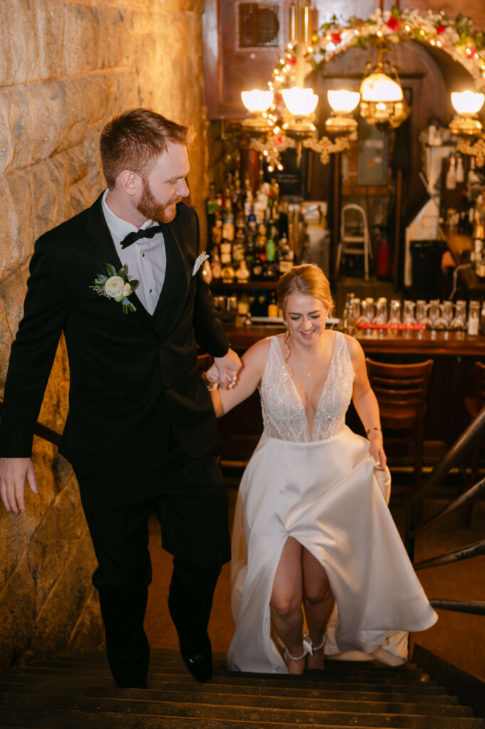 Bride and groom walking hand in hand up the staircase inside Union Station Banquets. Warm lighting, historic stone walls, and glowing bar details create a romantic and elegant wedding portrait with a vintage speakeasy atmosphere. Photographed by Boston area wedding Photographer