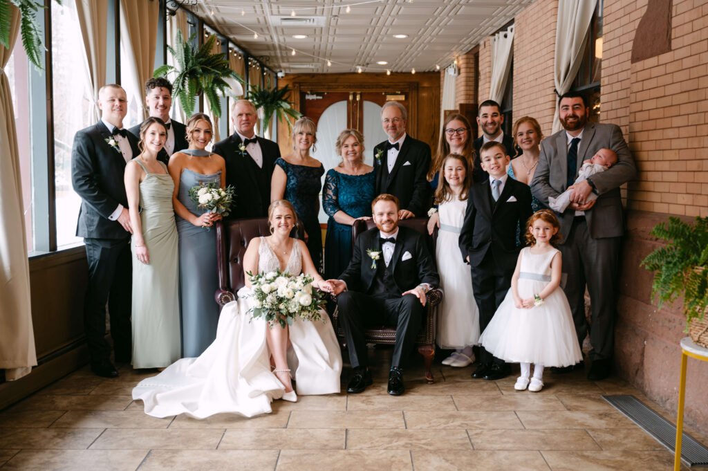 Bride and groom seated in leather chairs surrounded by family members during an indoor wedding portrait at Union Station Banquets. Guests and children are gathered around the couple in formal attire, with large windows, brick walls, greenery, and warm natural light creating an elegant and timeless atmosphere.
