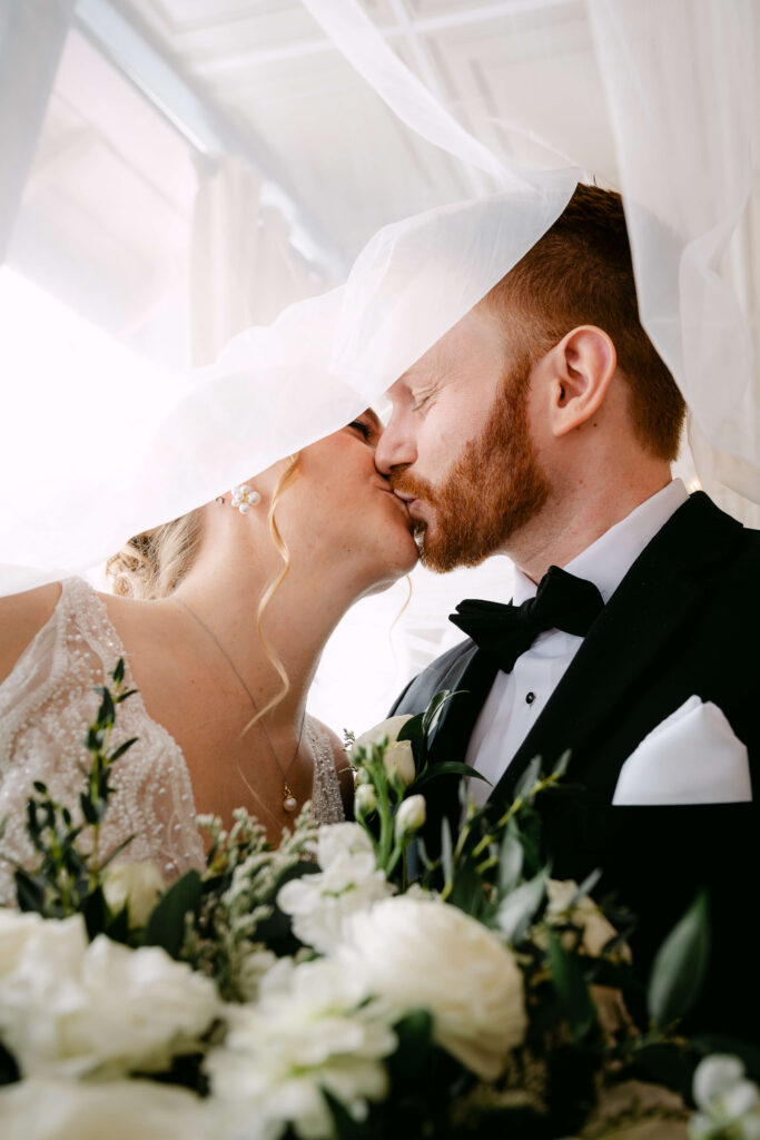 Bride and groom sharing a kiss beneath the bride’s veil during an intimate wedding portrait at Union Station Banquets. Soft natural light filters through the veil while white florals and greenery frame the foreground, creating a romantic and timeless moment.